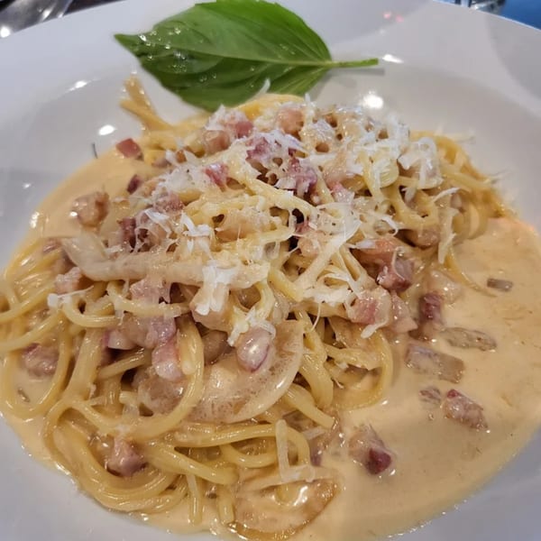 Creamy carbonara pasta with bacon bits, grated cheese, and a fresh basil leaf on a white plate.