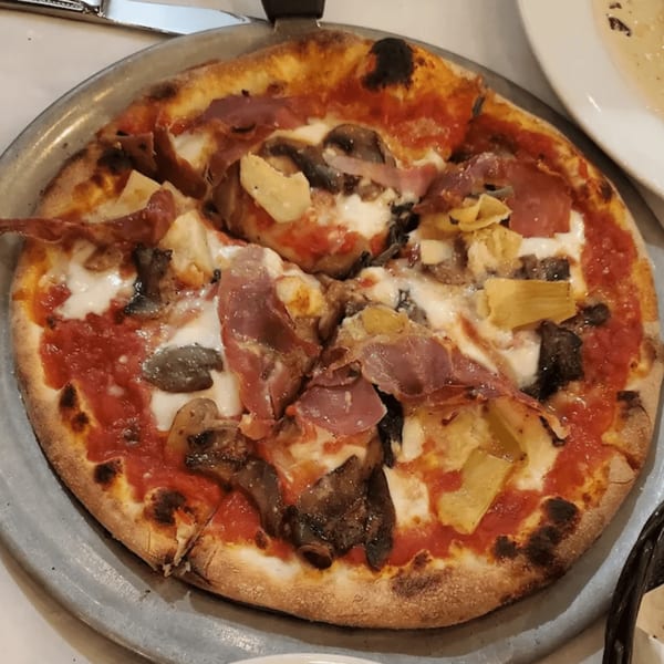 A whole Italian pizza with prosciutto, mushrooms, and artichoke hearts on a metal tray.
